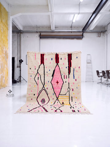A large Moroccan-style woven rug with a cream base and abstract geometric patterns in pink, black, red, yellow, and blue, hanging in a bright white studio space.